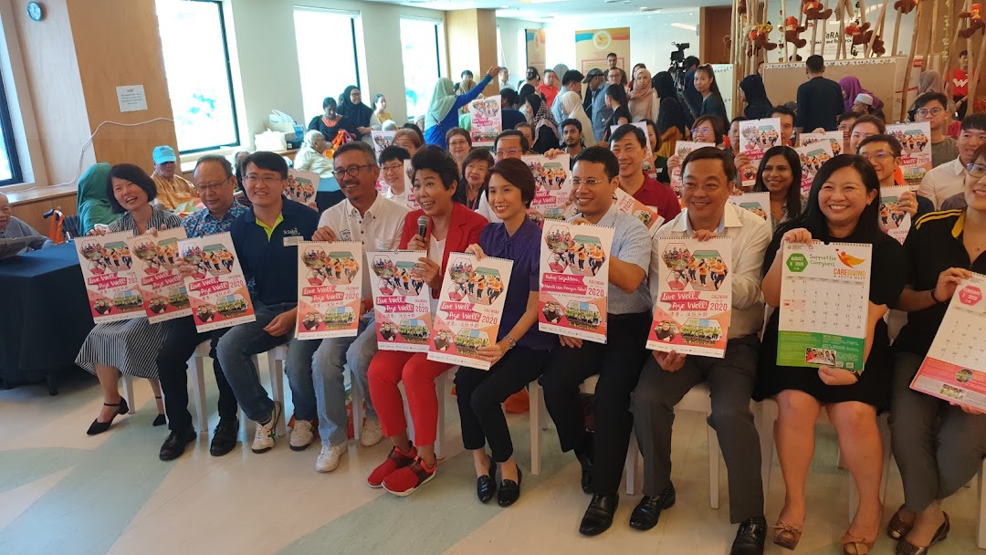 A group of smiling people, including community leaders, holding 'Live Well! Age Well! 2020' calendars at an indoor event.
