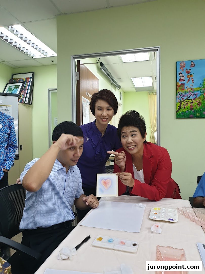 Three individuals engaging in an art therapy session, with one proudly displaying a heart painting.