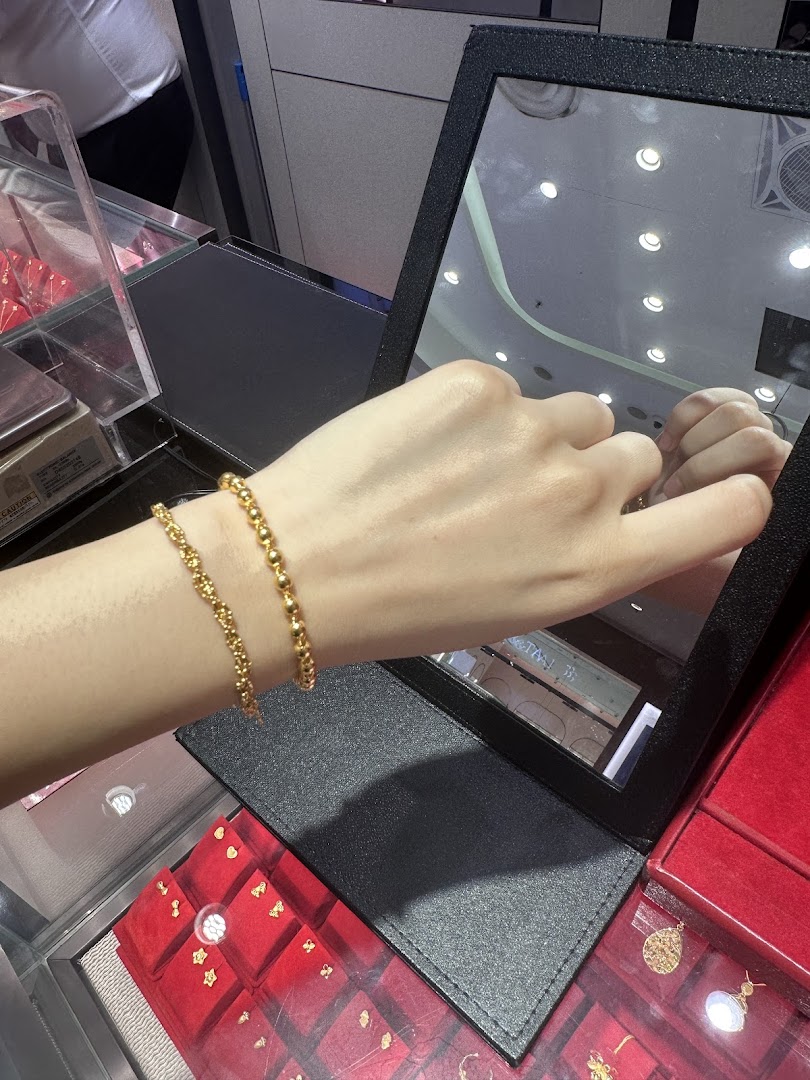 A hand models two delicate gold bead bracelets in a jewelry store, reflected in a mirror.