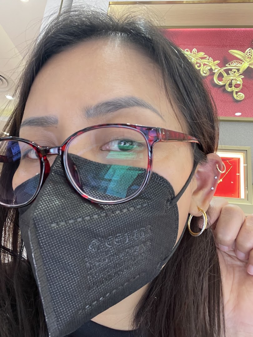 A person wearing a mask and glasses showcases a gold hoop earring, with gold jewelry displays in the background.