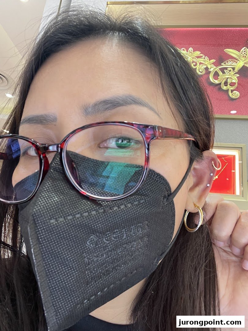A person wearing a mask and glasses showcases a gold hoop earring, with gold jewelry displays in the background.