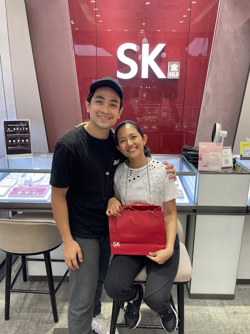 Two smiling customers pose inside SK Jewellery, holding a red shopping bag, with display cases visible.