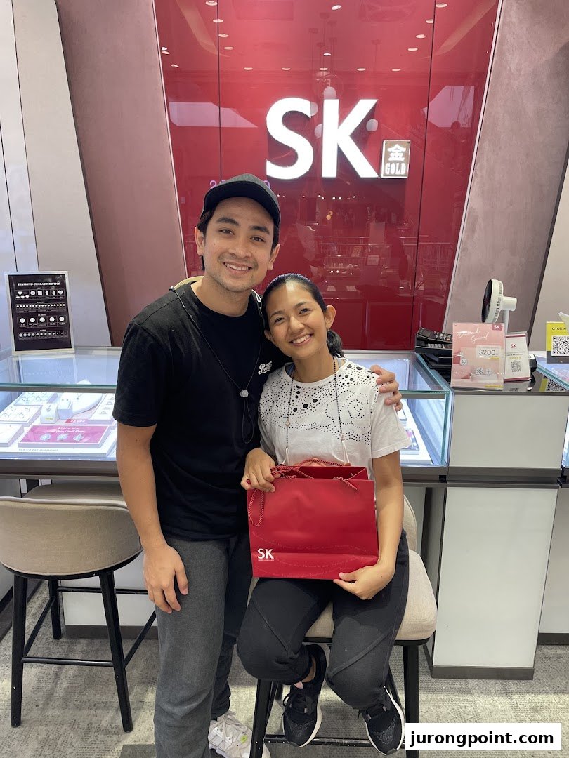Two smiling customers pose inside SK Jewellery, holding a red shopping bag, with display cases visible.