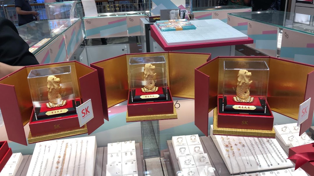 Three golden Merlion statues are prominently displayed in red boxes, surrounded by various jewelry items in a retail setting.