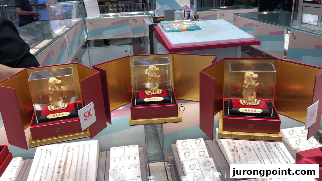 Three golden Merlion statues are prominently displayed in red boxes, surrounded by various jewelry items in a retail setting.