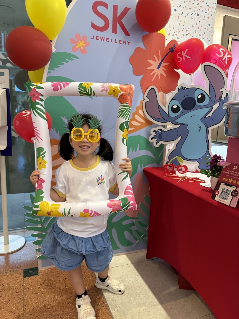 A child poses with a tropical-themed photo frame at an SK Jewellery event booth featuring Stitch.
