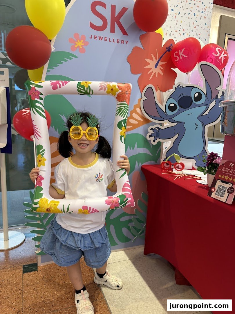 A child poses with a tropical-themed photo frame at an SK Jewellery event booth featuring Stitch.