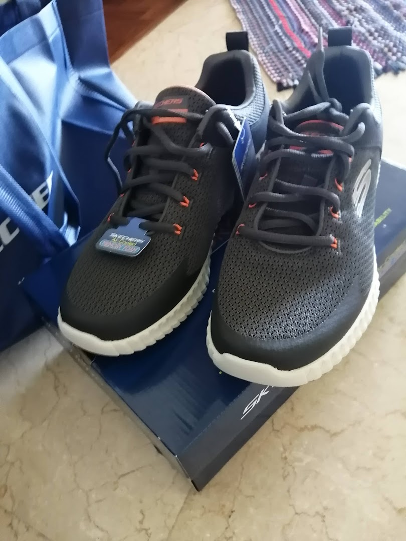 A new pair of dark grey Skechers sneakers with orange accents, resting on their box next to a blue shopping bag.