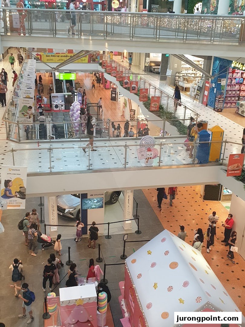 A vibrant multi-level shopping mall interior bustling with shoppers, various stores, and promotional displays.