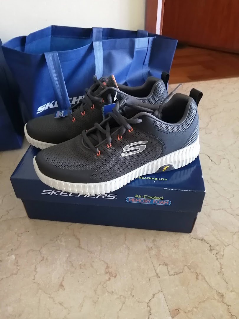 A pair of dark grey Skechers shoes with white soles on their box, next to a blue Skechers shopping bag.
