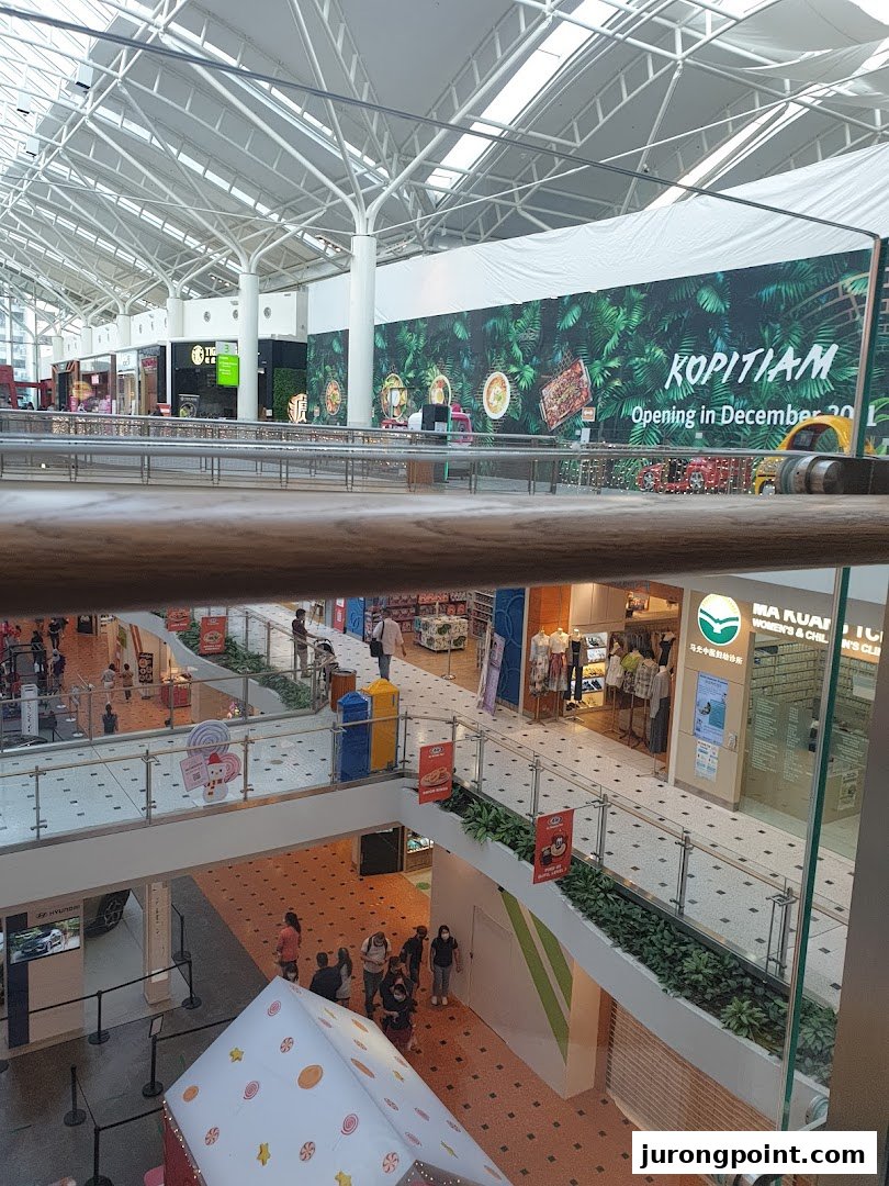 A multi-level mall interior with a high ceiling, showing various shops, a Kopitiam banner, and shoppers.