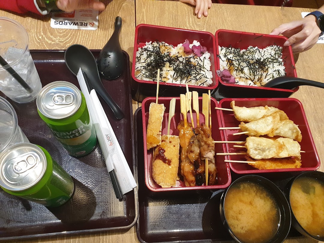 A top-down view of a meal featuring bento boxes with rice, skewers, fried items, miso soup, and canned drinks.