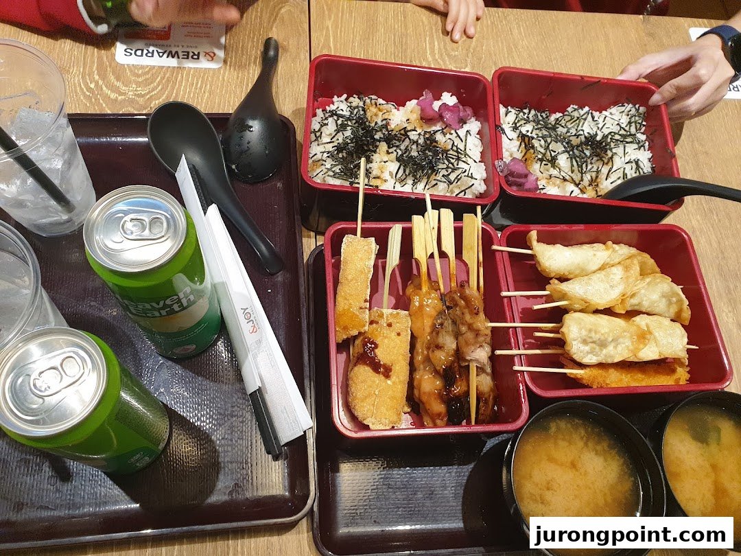 A top-down view of a meal featuring bento boxes with rice, skewers, fried items, miso soup, and canned drinks.