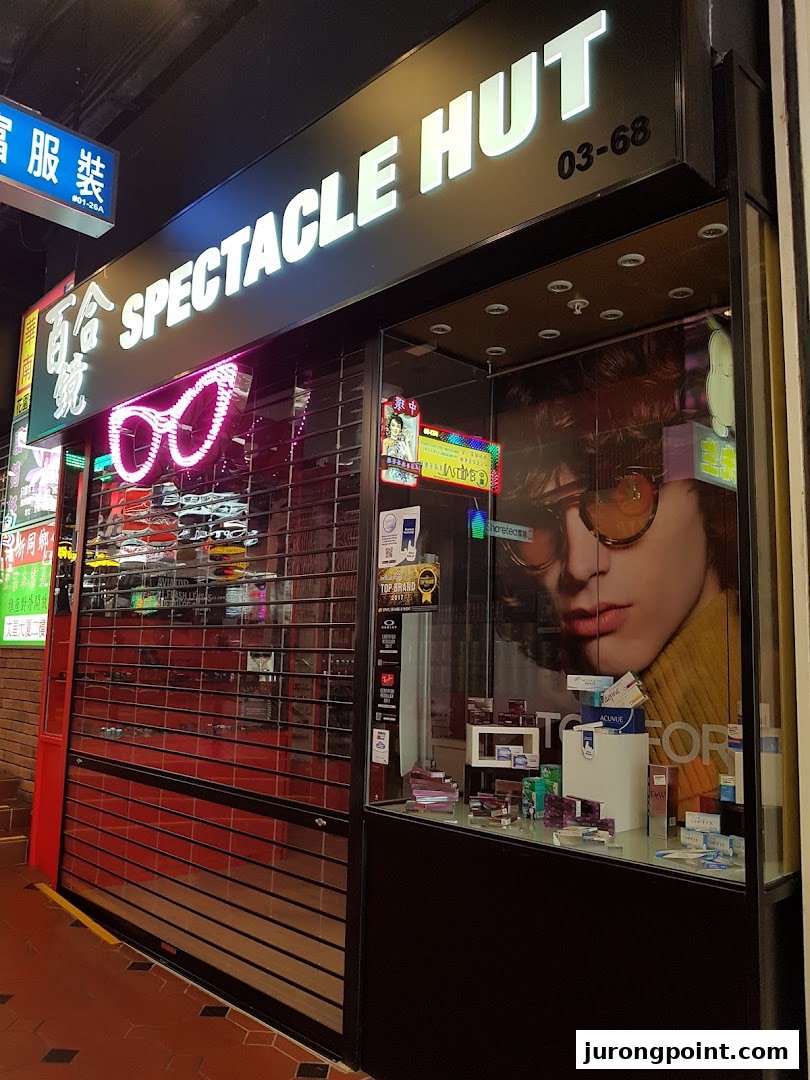 Illuminated storefront of Spectacle Hut, featuring a display window with eyewear and a closed security shutter.