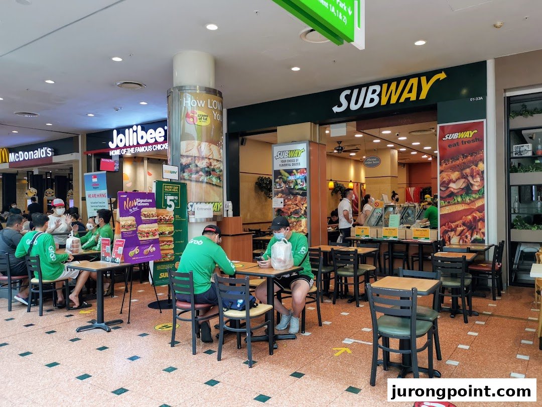 Subway shop interior with customers dining, showing its entrance and adjacent fast-food restaurants in a mall.
