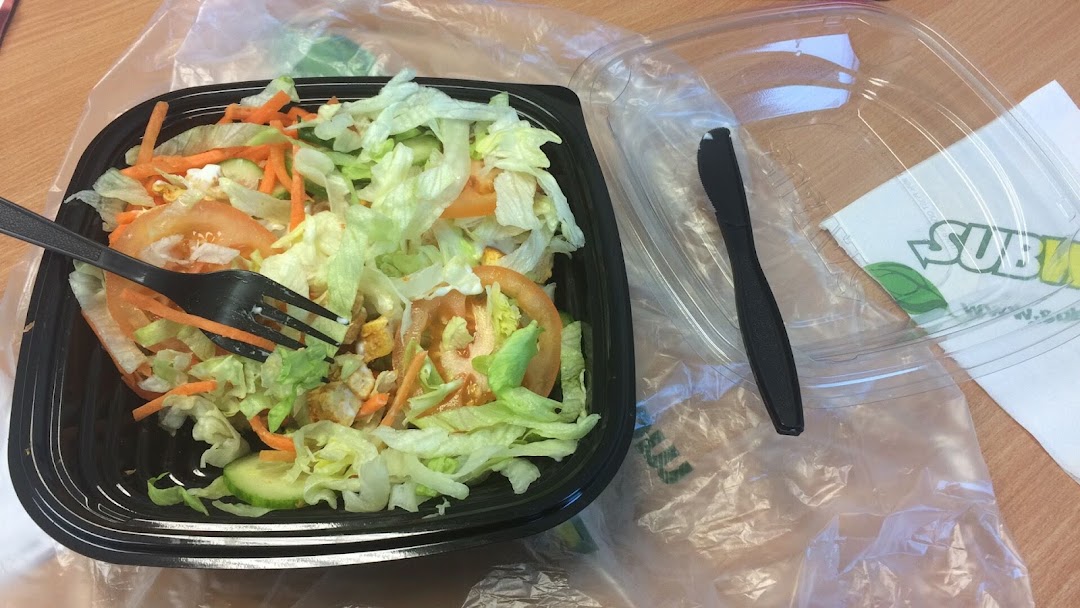 A fresh Subway salad in a black container with a fork, alongside a plastic lid and Subway napkin.