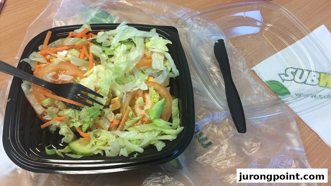 A fresh Subway salad in a black container with a fork, alongside a plastic lid and Subway napkin.