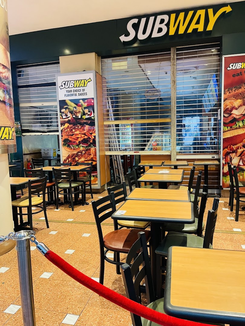Subway shop entrance with closed rolling shutter, outdoor seating, and promotional posters.