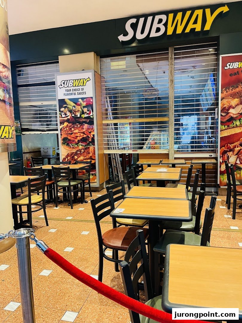 Subway shop entrance with closed rolling shutter, outdoor seating, and promotional posters.