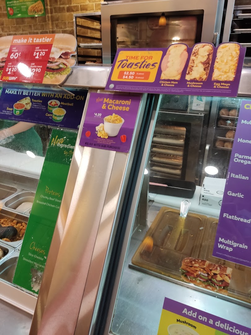 Subway counter displaying fresh ingredients, promotional offers for toasties, macaroni, and add-ons.