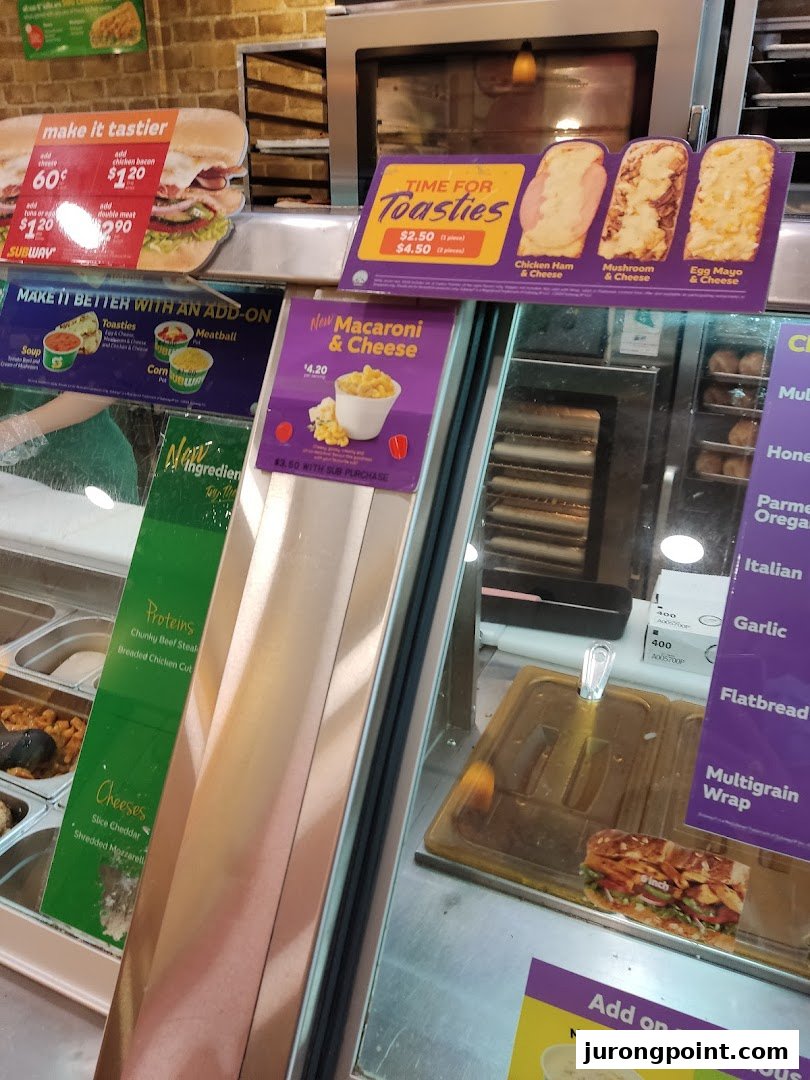 Subway counter displaying fresh ingredients, promotional offers for toasties, macaroni, and add-ons.