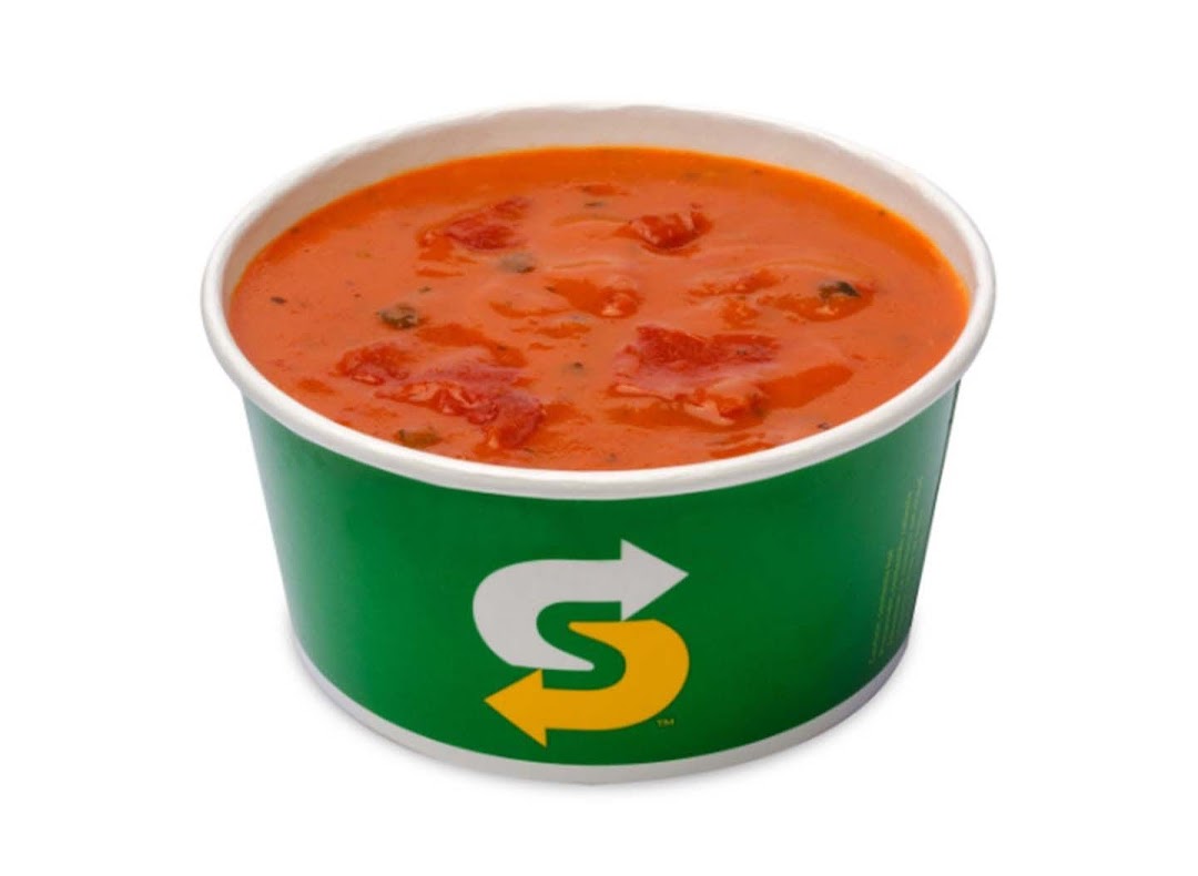 A vibrant orange tomato soup in a green Subway branded cup on a white background.
