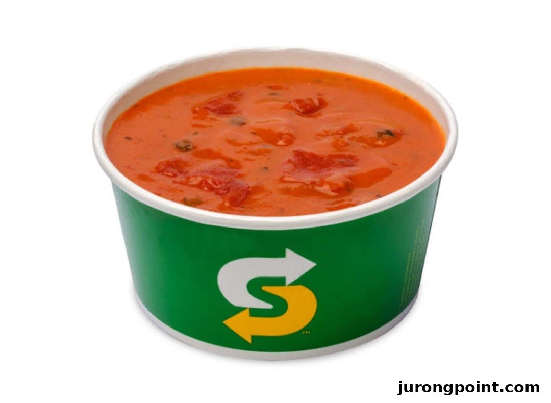 A vibrant orange tomato soup in a green Subway branded cup on a white background.