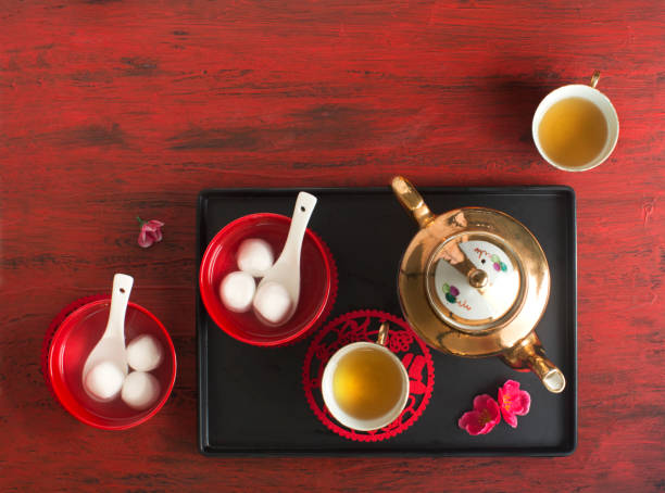 Overhead view of traditional Chinese tangyuan and a golden tea set on a black tray against a red background.