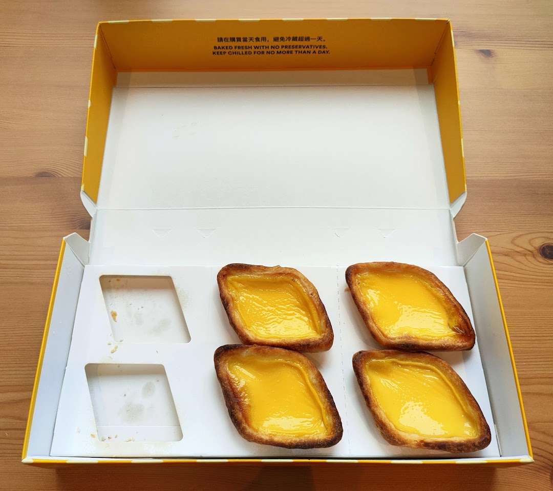 Four golden egg tarts in a yellow and white box, with two empty slots, on a wooden surface.