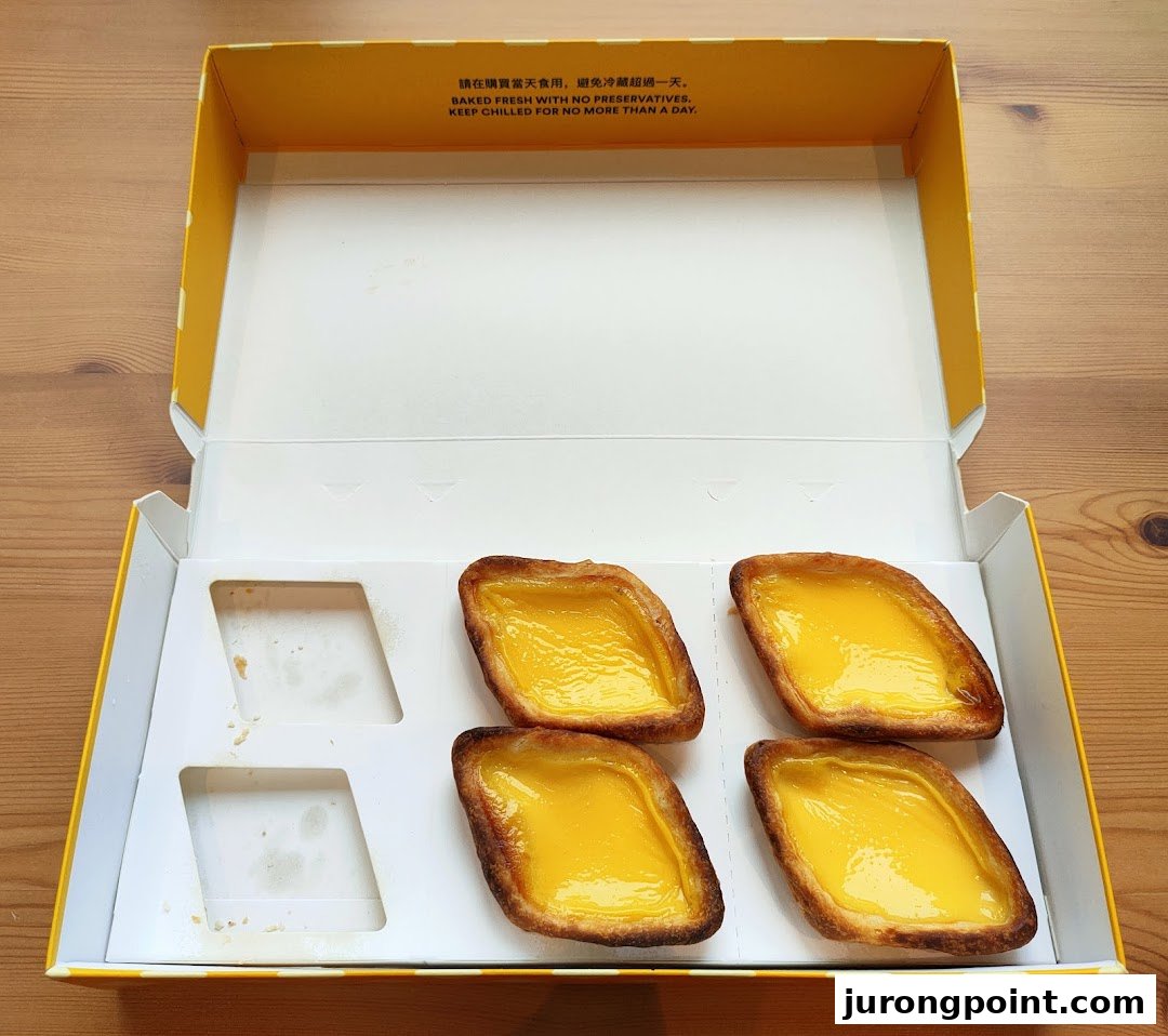 Four golden egg tarts in a yellow and white box, with two empty slots, on a wooden surface.
