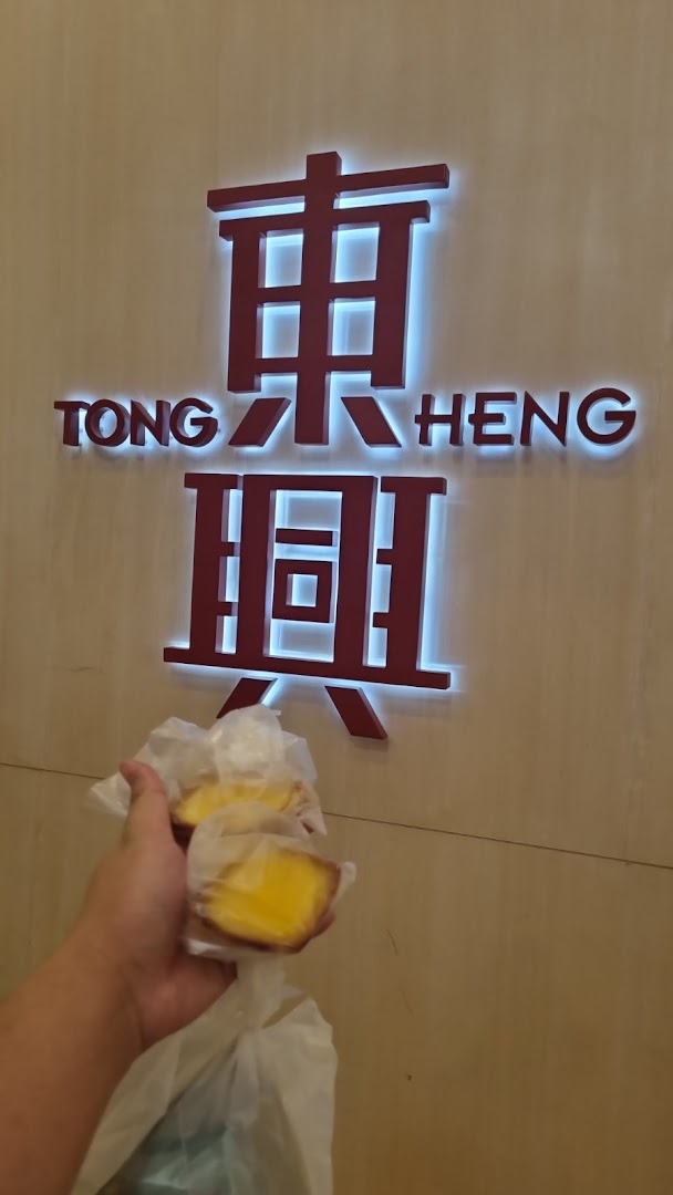 A hand holds two golden egg tarts in front of the illuminated Tong Heng bakery sign.