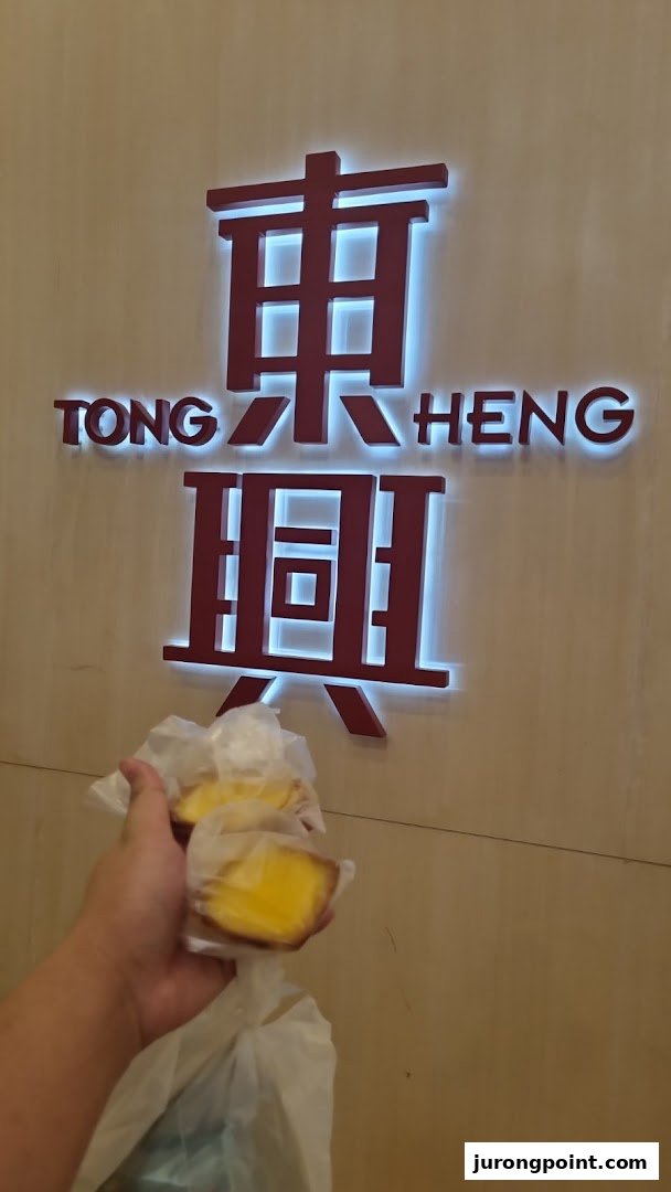 A hand holds two golden egg tarts in front of the illuminated Tong Heng bakery sign.