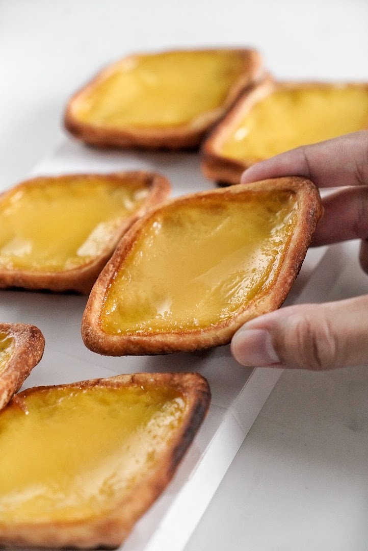 A hand picks up a golden diamond-shaped egg tart from a tray of several pastries.
