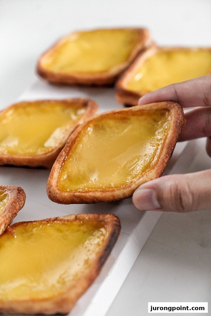 A hand picks up a golden diamond-shaped egg tart from a tray of several pastries.