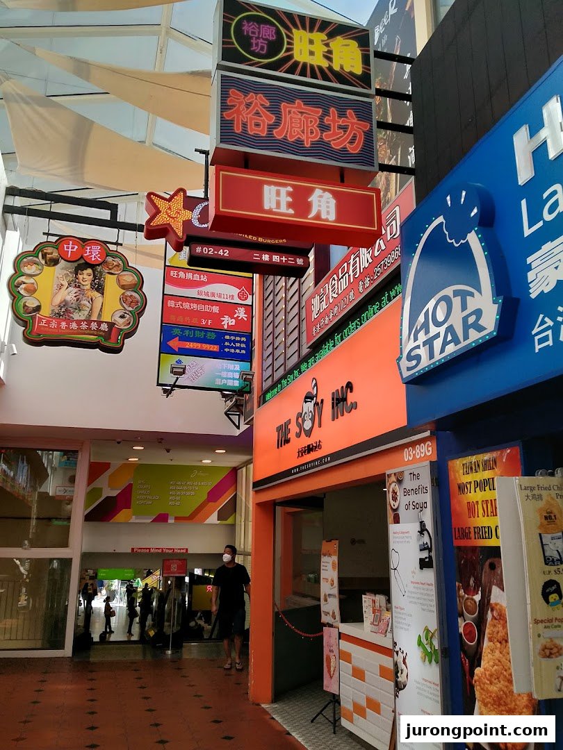 A vibrant mall walkway featuring diverse shop fronts including The Soy Inc. and Hot Star Large Fried Chicken.