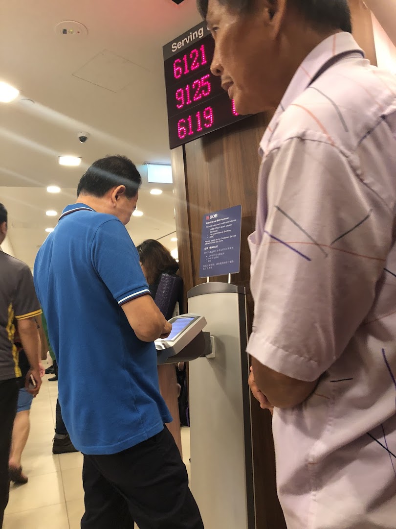 Customers are seen inside a UOB bank branch, with a digital queue display showing serving numbers.