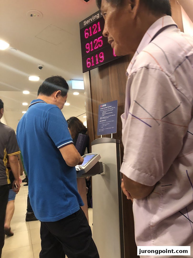 Customers are seen inside a UOB bank branch, with a digital queue display showing serving numbers.