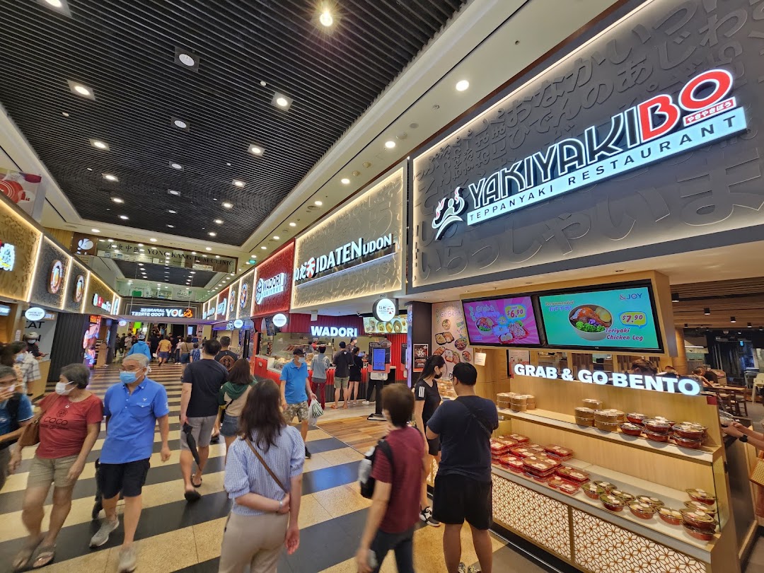 A bustling mall food court featuring various Japanese restaurants like Yakiyakibo and Idaten Udon, with many shoppers.
