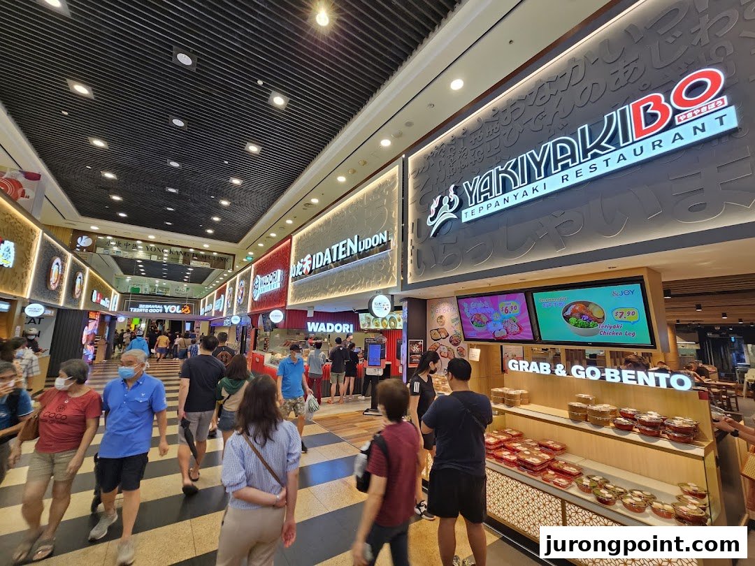 A bustling mall food court featuring various Japanese restaurants like Yakiyakibo and Idaten Udon, with many shoppers.