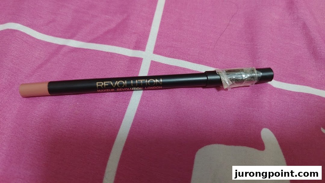 A black Makeup Revolution London pencil with a nude tip on a pink patterned surface.