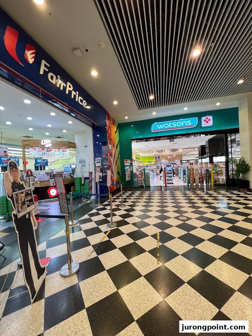Entrance to Watsons pharmacy and FairPrice supermarket from a mall corridor with a checkered floor.