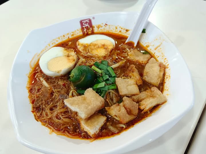 A close-up of a delicious bowl of spicy noodles with eggs, lime, and crispy toppings from Ya Kun Family Cafe.