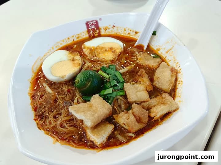 A close-up of a delicious bowl of spicy noodles with eggs, lime, and crispy toppings from Ya Kun Family Cafe.