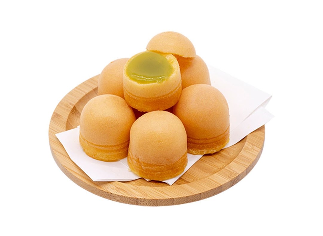 A wooden plate holds several golden-brown pastries, one revealing a vibrant green pandan kaya filling.