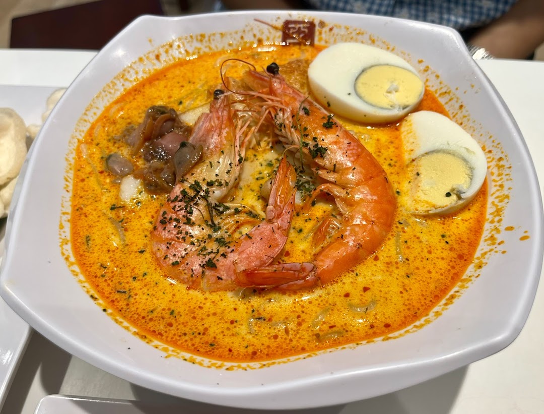 A vibrant bowl of spicy noodle soup with large prawns, clams, and hard-boiled eggs.