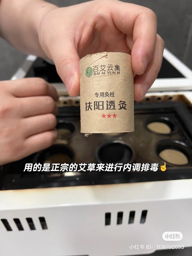 A hand holds a moxa stick over a moxibustion device, highlighting a TCM treatment.