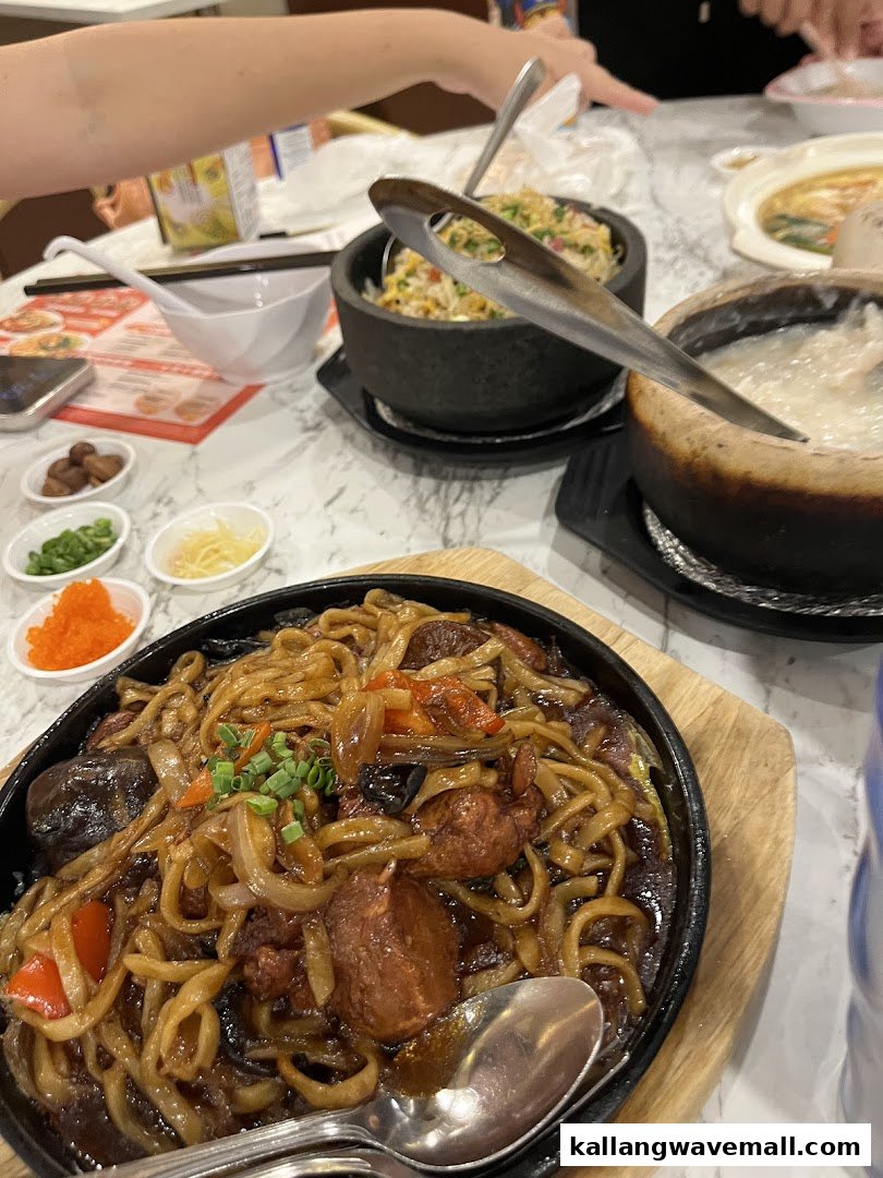 A close-up of a sizzling plate of noodles with meat and vegetables, alongside other dishes like fried rice and porridge.