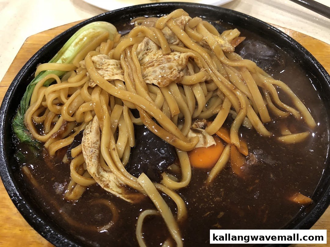 A close-up of savory noodles with vegetables and sauce served on a sizzling hot plate.