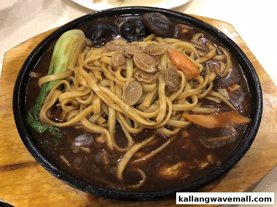 A sizzling plate of noodles with abalone slices, bok choy, carrots, and mushrooms in a rich sauce.