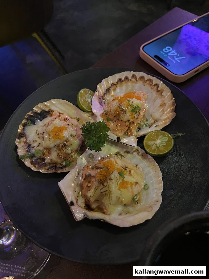 A plate of three baked scallops topped with cheese, roe, and lime wedges.